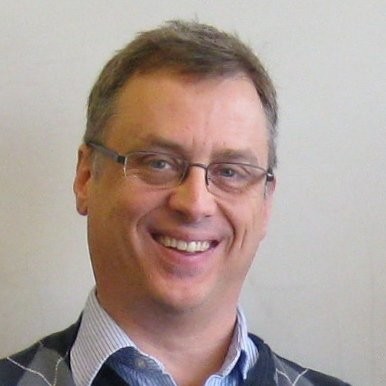 Smiling man wearing glasses and a light blue striped shirt with dark sweater on top