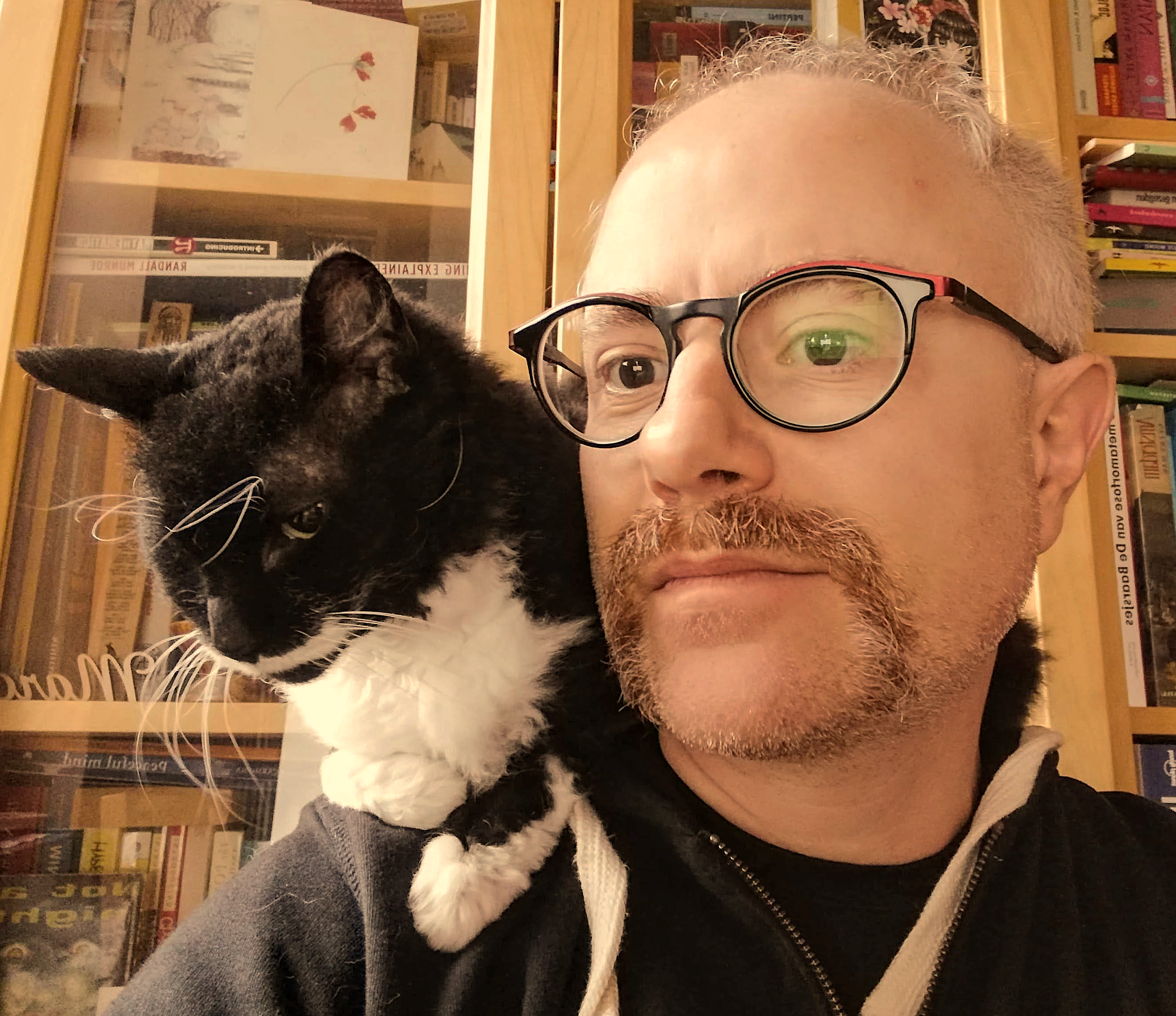 a man with a cat on his shoulder standing in front of a book cabinet